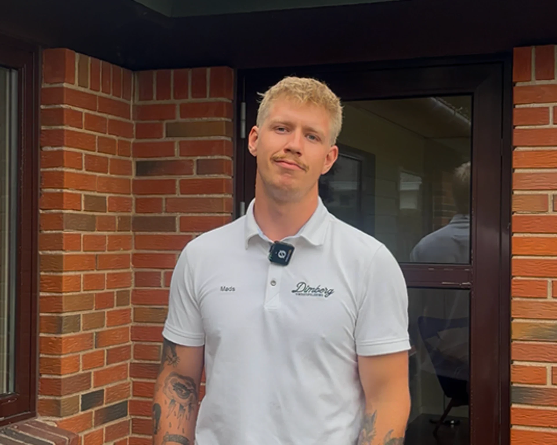 Mand i hvid polo med Dimberg ApS logo foran en murstensbygning.